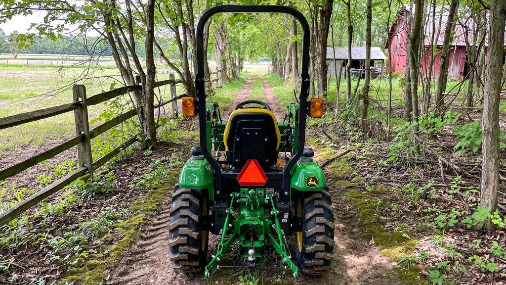 John Deere 3038E maneuvering on a narrow trail between trees on a small farm